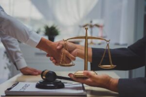 Two people shaking hands in a legal office with a scale of justice and gavel on the desk, symbolizing agreement and legal support