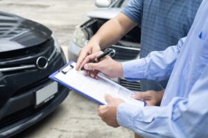 insurance officer writing on clipboard while insurance agent examining black car after accident
