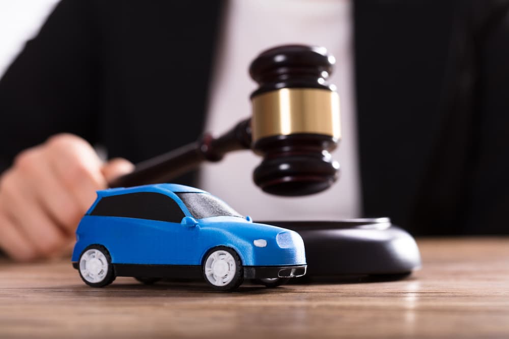 A tiny blue car stands before a judge, who holds a gavel in hand.