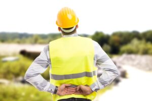 Construction worker in a yellow safety vest and hard hat holding his lower back in pain at a construction site.