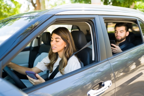 Young female rideshare driver looking at her phone while driving, with a male passenger in the back seat using his own phone.