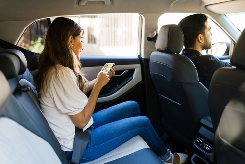 Woman sitting in the backseat of a car wearing a seatbelt and using her smartphone during a rideshare trip.