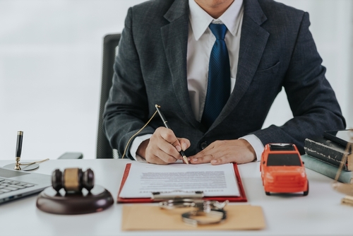 Lawyer in a suit reviewing and signing legal documents at a desk with a gavel, small car model, and handcuffs symbolizing an auto accident case.