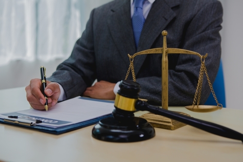 A lawyer in a suit writes on a clipboard next to a golden balance scale and a gavel, representing personal injury legal work and documentation.