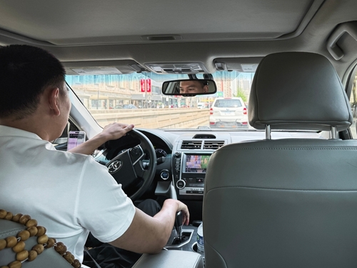 View from the back seat of a car showing a male driver wearing a white shirt and wooden bead bracelet while driving through city traffic.