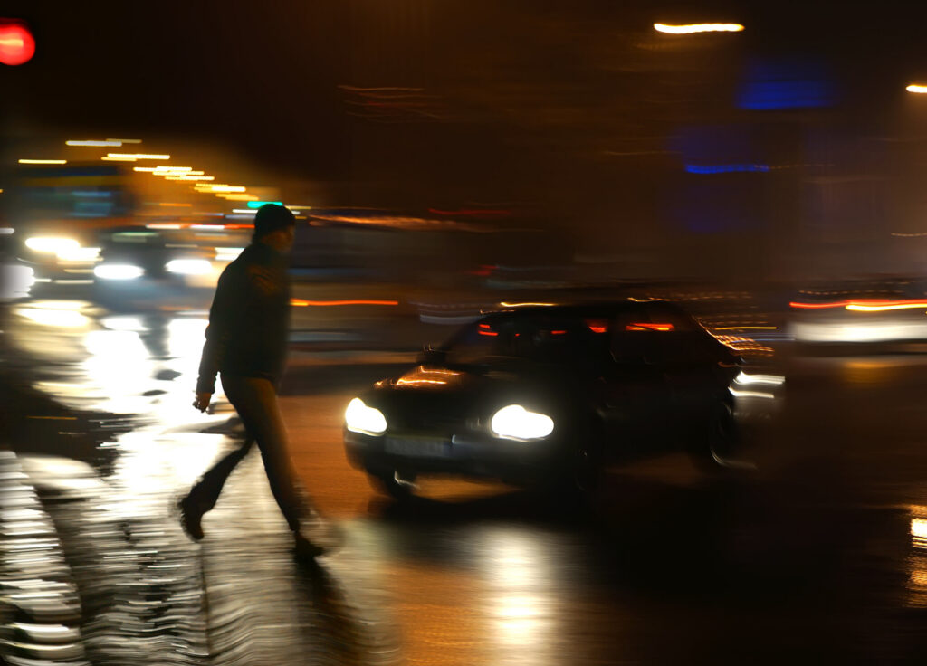 Un coche, con el movimiento difuminado, se aproxima a un peatón que cruza una calle oscura, lo que ilustra los peligros de los accidentes de peatones en Bakersfield.