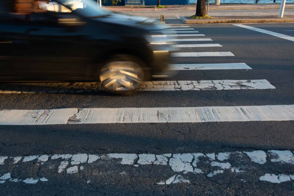 A blurred car driving across a white-striped crosswalk in Bakersfield, representing the shared duty of care between drivers and pedestrians.