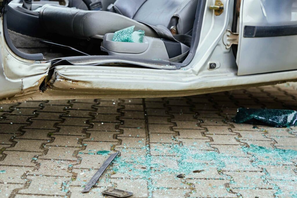 Green shattered glass scattered on paving stones next to a severely damaged vehicle with an open door and deployed airbags, showing a severe wreck scene handled by Bakersfield catastrophic injury lawyers.