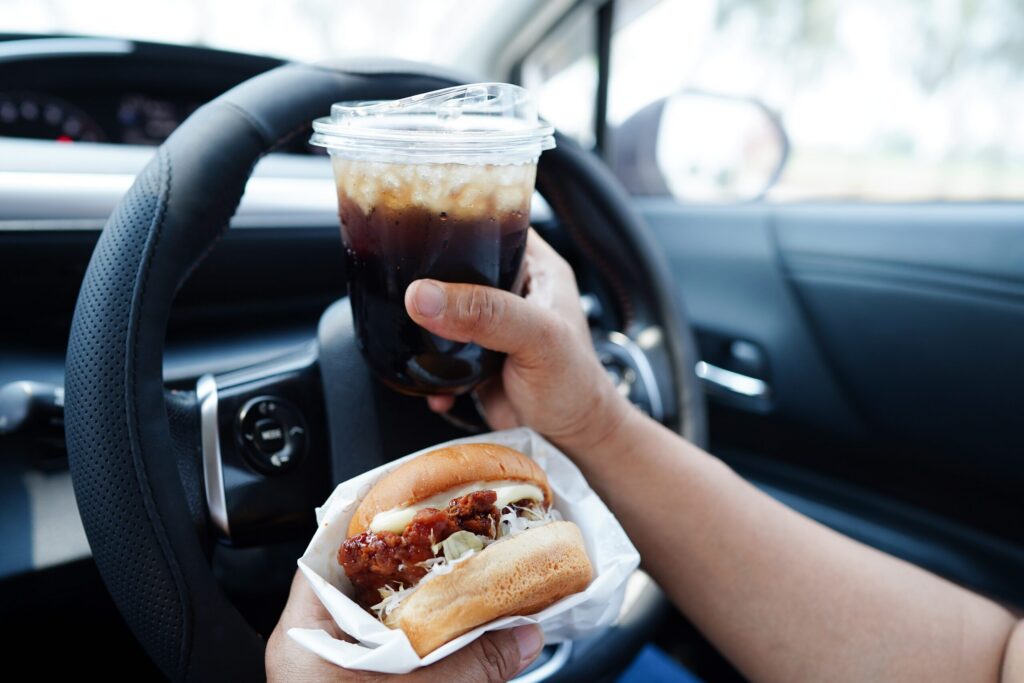A driver holds a chicken burger in one hand and an iced coffee in the other while behind the steering wheel, showcasing a dangerous behavior handled by Bakersfield distracted driving accident lawyers.