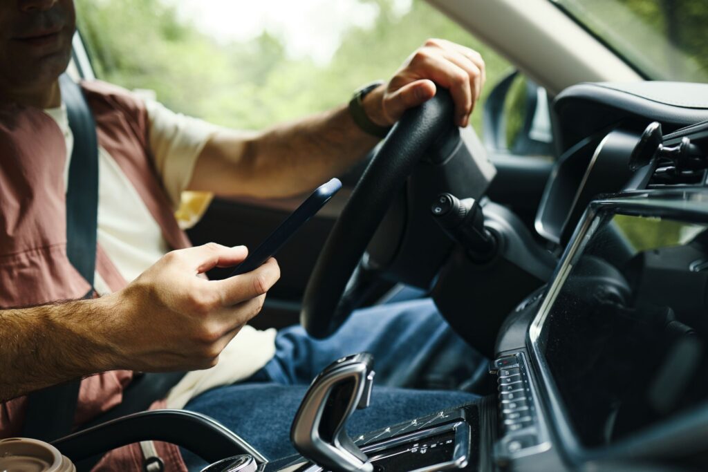 A man uses his smartphone with one hand while keeping his other hand on the steering wheel while driving, illustrating a negligent action investigated by Bakersfield distracted driving accident lawyers.