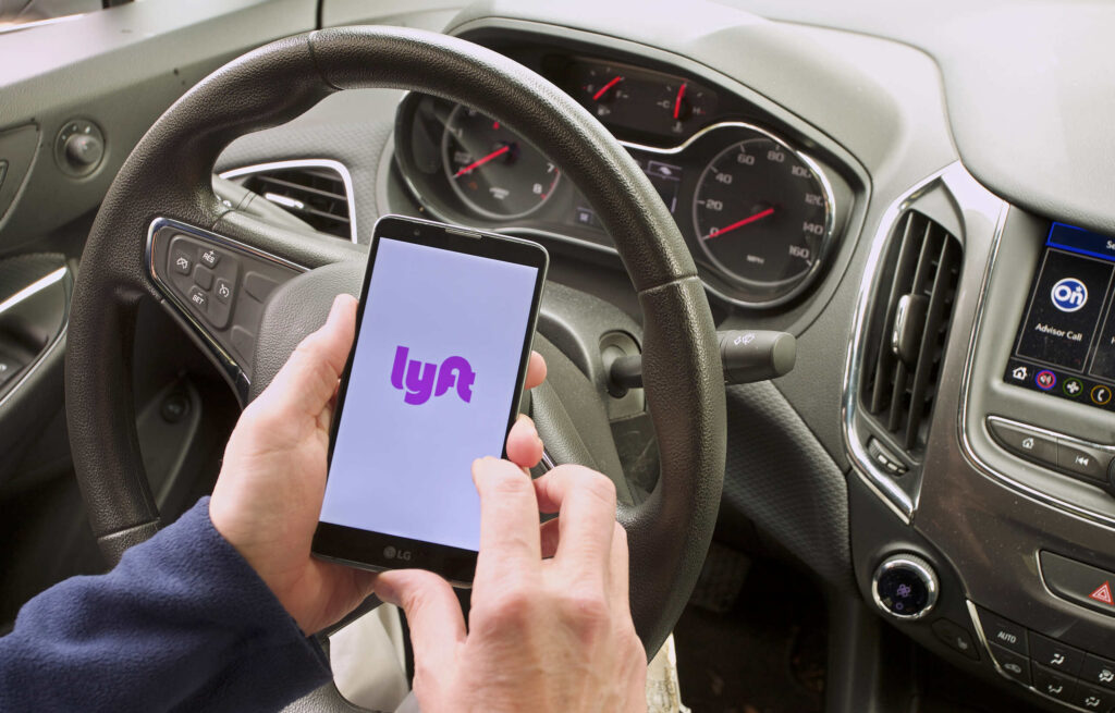 A driver holds a smartphone displaying the pink Lyft logo while sitting behind the steering wheel inside a vehicle, highlighting rideshare app usage analyzed by Bakersfield Lyft accident lawyers.