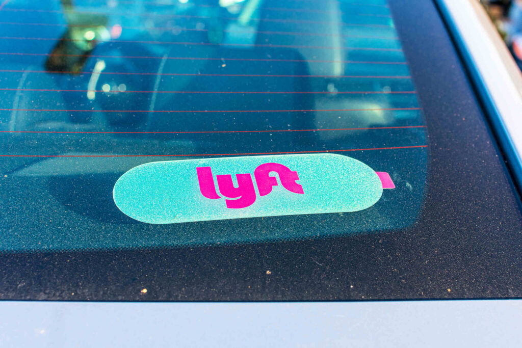 A driver holds a smartphone displaying the pink Lyft logo while sitting behind the steering wheel inside a vehicle, highlighting rideshare app usage analyzed by Bakersfield Lyft accident lawyers.