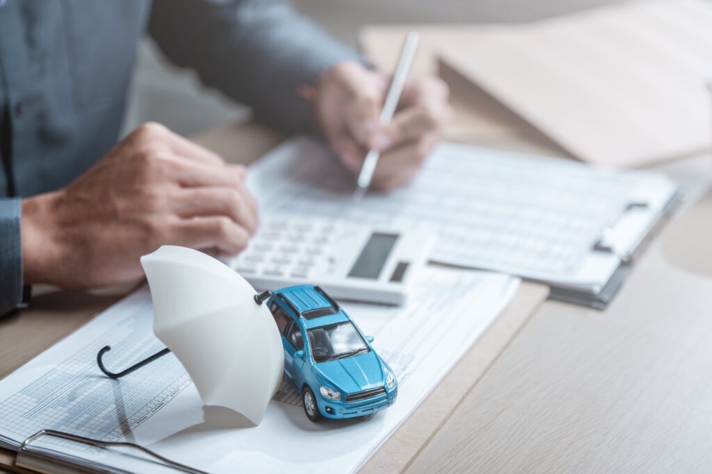 A small toy car sits under a white miniature umbrella on top of legal paperwork while a professional uses a calculator in the background, symbolizing coverage review by Bakersfield uninsured/underinsured motorist accident lawyers.