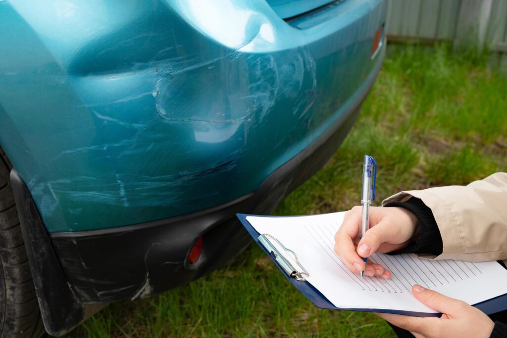 A person taking notes on a clipboard near a scratched blue bumper illustrates how long do you have to report a car accident to your insurance in California.