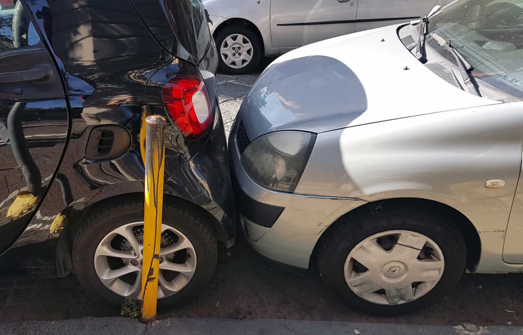 A silver car hitting a black car parked next to a yellow pole illustrates questions about Who's Liable for a Car Accident in a Bakersfield Parking Lot.