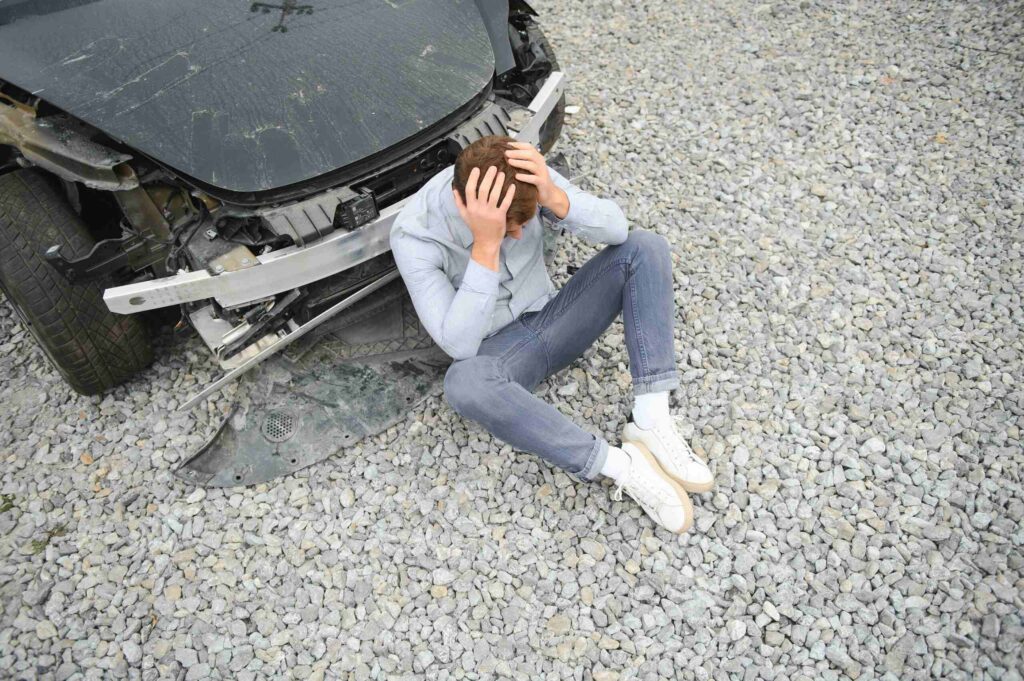 A distressed man holding his head while sitting on gravel next to a damaged car.