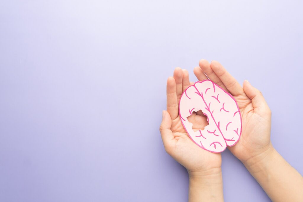 Two hands holding a paper cutout of a human brain with a missing puzzle piece.